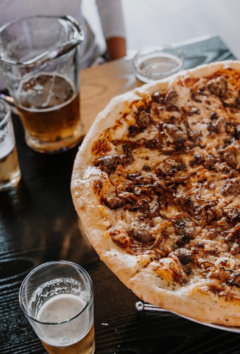 A large, hot pizza with meat and cheese, accompanied by several glasses of beer on a dark table.